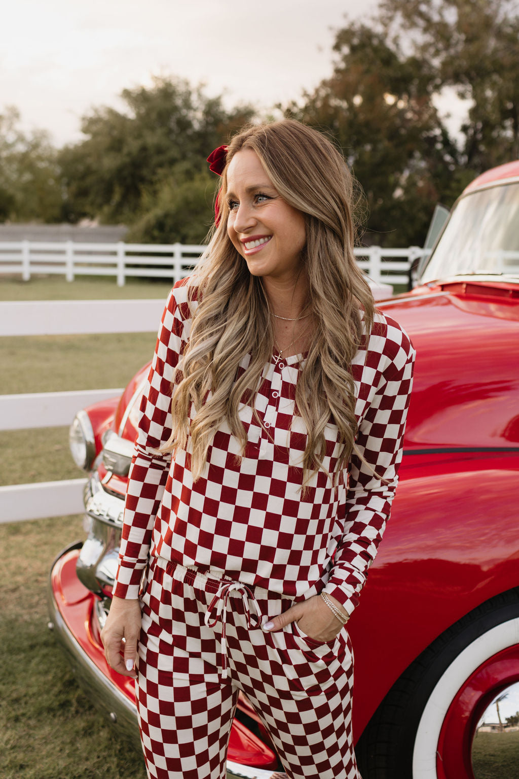 A person smiles while leaning against a vintage red and white truck, dressed in lolo webb's Women's Bamboo Pajamas | the Quinn, which complements their long, wavy blonde hair. The serene scene features a white fence and green grass in the background.