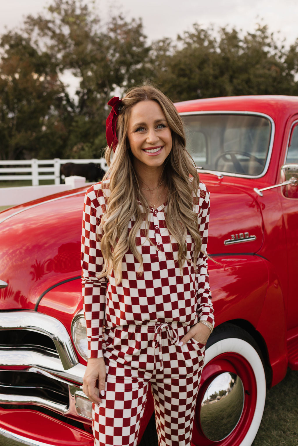 A woman, smiling in her lolo webb Women's Bamboo Pajamas | the Quinn, designed for comfort and style, stands in front of a vintage red truck. Trees are visible in the background, complementing her charming hypo-allergenic clothing.