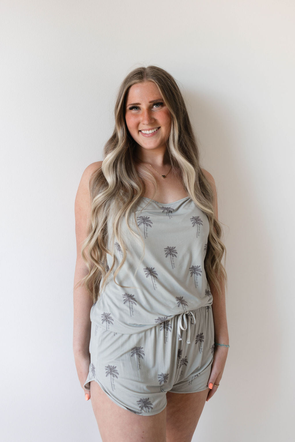 A woman with long, wavy blonde hair is smiling and standing against a plain white background. She is wearing the hypo-allergenic, light grey Cami Women's Bamboo Set | Summer Dreamin' with palm tree prints by Forever French.