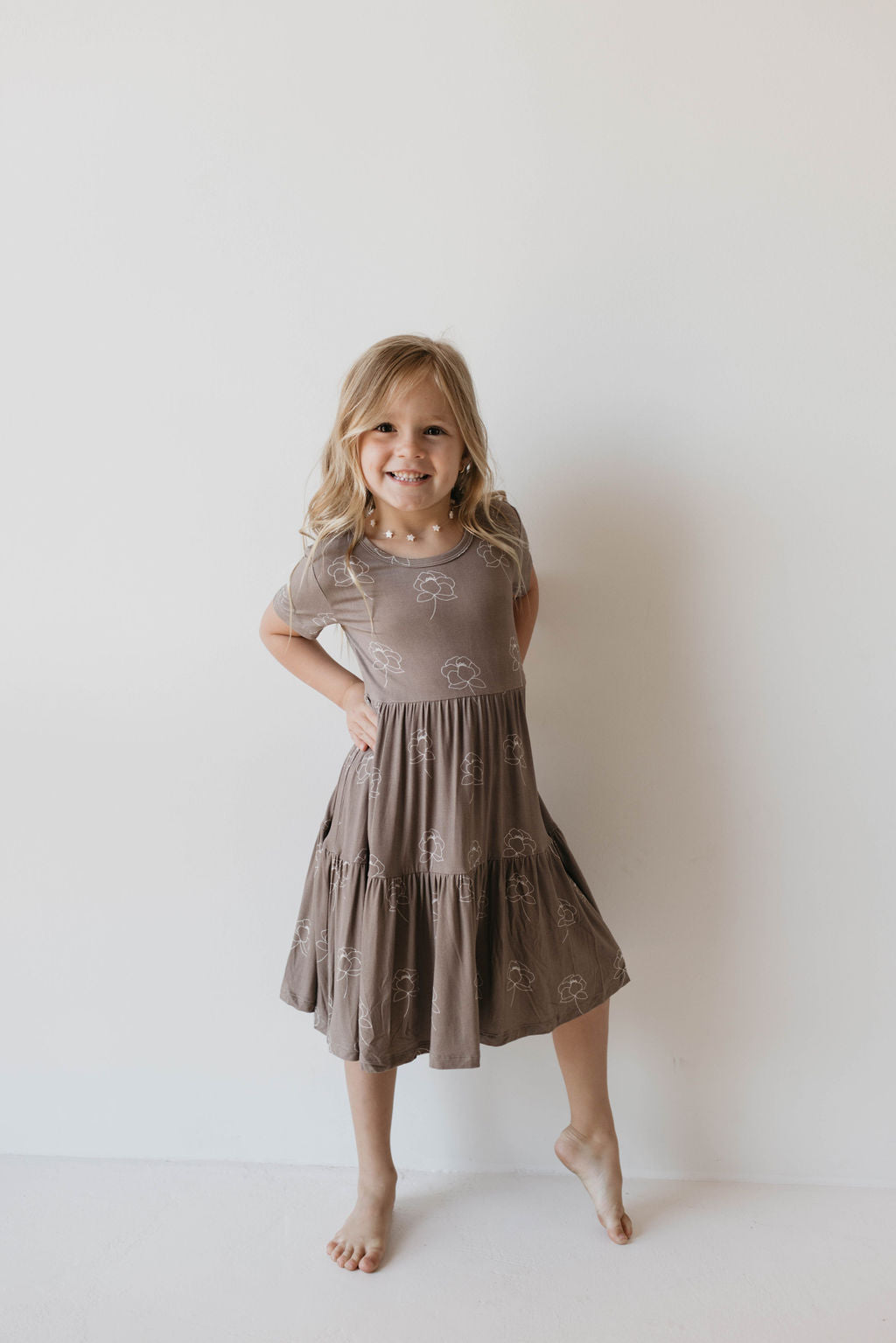 A young girl with long, light blonde hair smiles while standing against a plain white wall. She is wearing the knee-length, short-sleeved "Multi Tier Bamboo Dress | Sweet Dreams Floral" by forever french baby. One of her legs is slightly bent, and her hands rest on her hips.