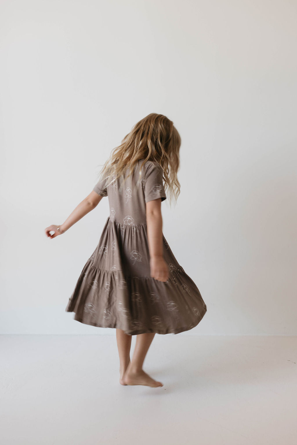 A young girl with long, wavy blonde hair spins around barefoot, wearing a knee-length, short-sleeved Multi Tier Bamboo Dress in the Sweet Dreams Floral pattern by Forever French Baby. The background is a plain, light-colored wall.