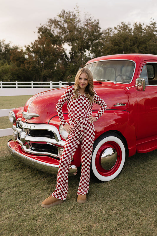 A young girl wearing the Pre-Teen Flare Bamboo Pajamas by lolo webb, characterized by their red and white checkered design, stands confidently in front of a classic red Chevrolet truck. She poses with a hand on her hip, while a white fence and trees grace the background, adding charm to the serene scene.