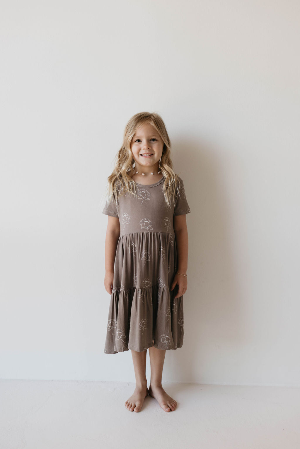 A young girl stands barefoot against a plain white wall, smiling. She is dressed in the "Multi Tier Bamboo Dress | Sweet Dreams Floral" by forever french baby. Her long, wavy blonde hair cascades past her shoulders as she rests her hands at her sides.