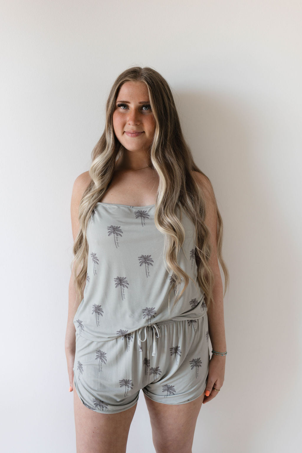 A woman with long, wavy hair dons the Forever French Cami Women's Bamboo Set in Summer Dreamin', featuring a charming light gray romper with a palm tree print. Made from hypo-allergenic bamboo fabric, she stands against a plain white background with her hands by her sides.