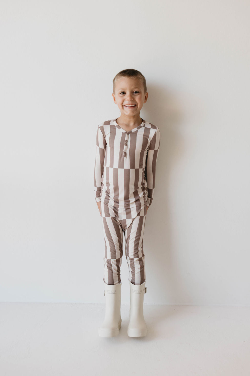 A young child stands against a plain white background, smiling. They are wearing cozy sleepwear—a long-sleeved, beige and white striped Bamboo Two Piece Pajama set from forever french baby, along with white rain boots. The child has short hair and their hands rest casually by their sides, showcasing the charm of Forever French Baby fashion.