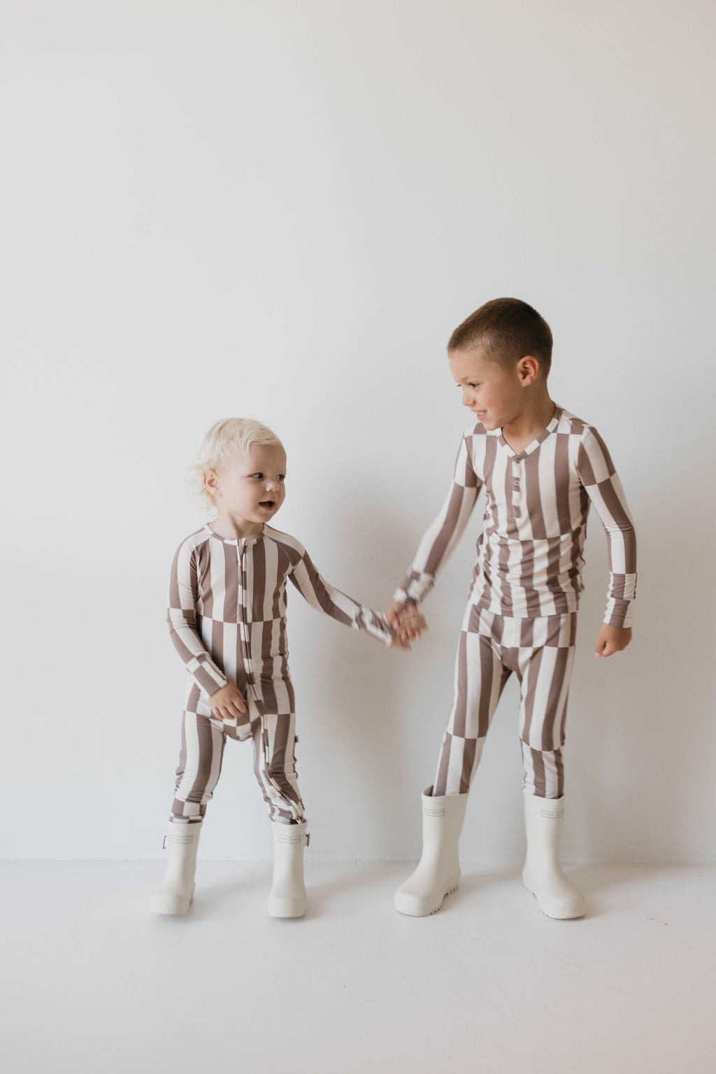 Two young children are wearing matching Bamboo Zip Pajamas in the Tile Stack pattern by forever french baby, along with white boots. The smaller child has blonde hair, while the older child has short brown hair. They are standing against a plain white background, holding hands and smiling at each other.