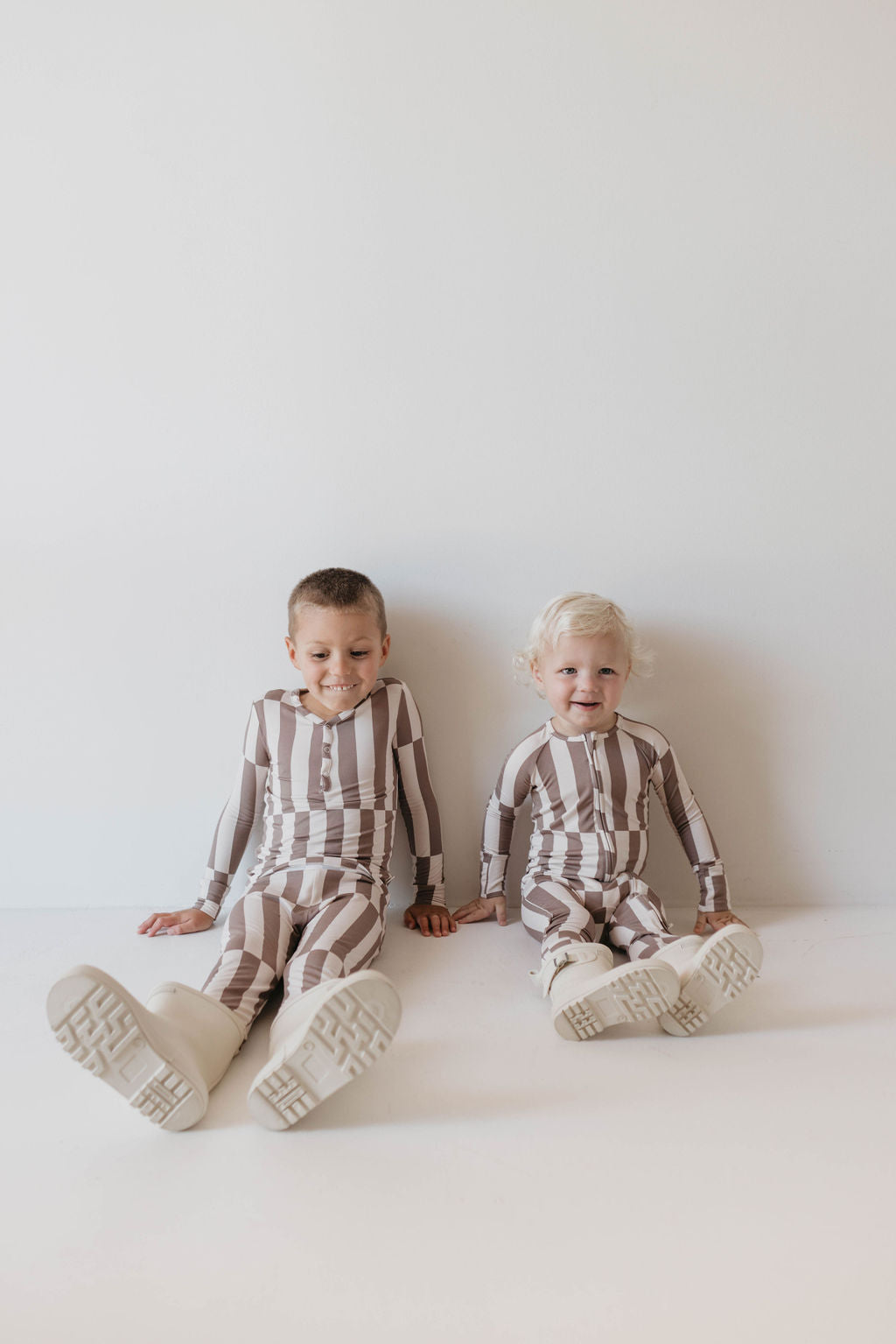 Two young children sit against a white wall, smiling in their cozy Bamboo Two Piece Pajamas | Tile Stack from Forever French Baby. They wear matching long-sleeved, striped pajamas and white shoes. The child on the left has short, dark hair, while the child on the right has short, blonde hair.