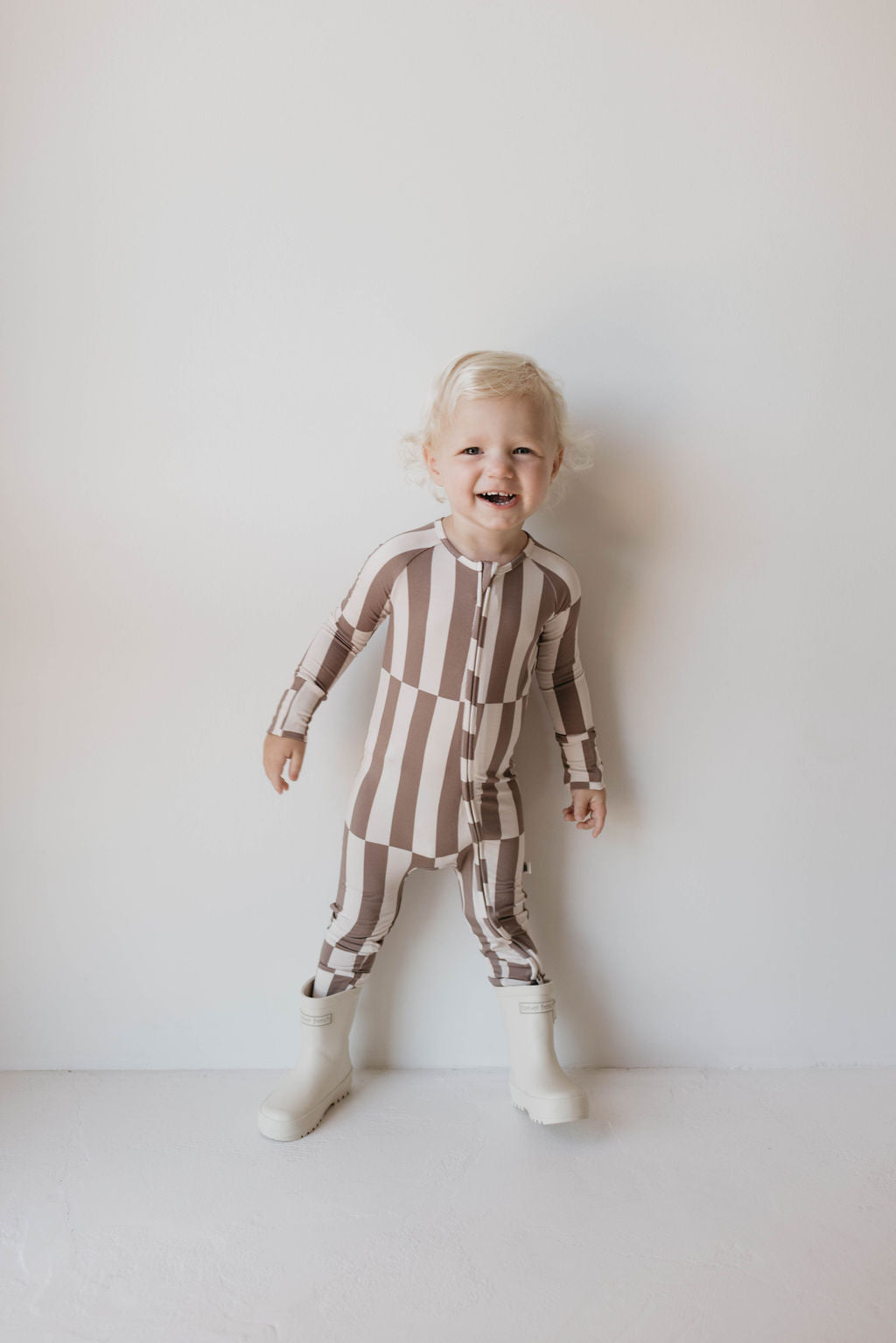 A small child with curly blonde hair smiles while standing against a plain background. They are slightly turned to the side, with hands by their sides. The child is wearing Forever French Baby Bamboo Zip Pajamas in the Tile Stack pattern and white boots.