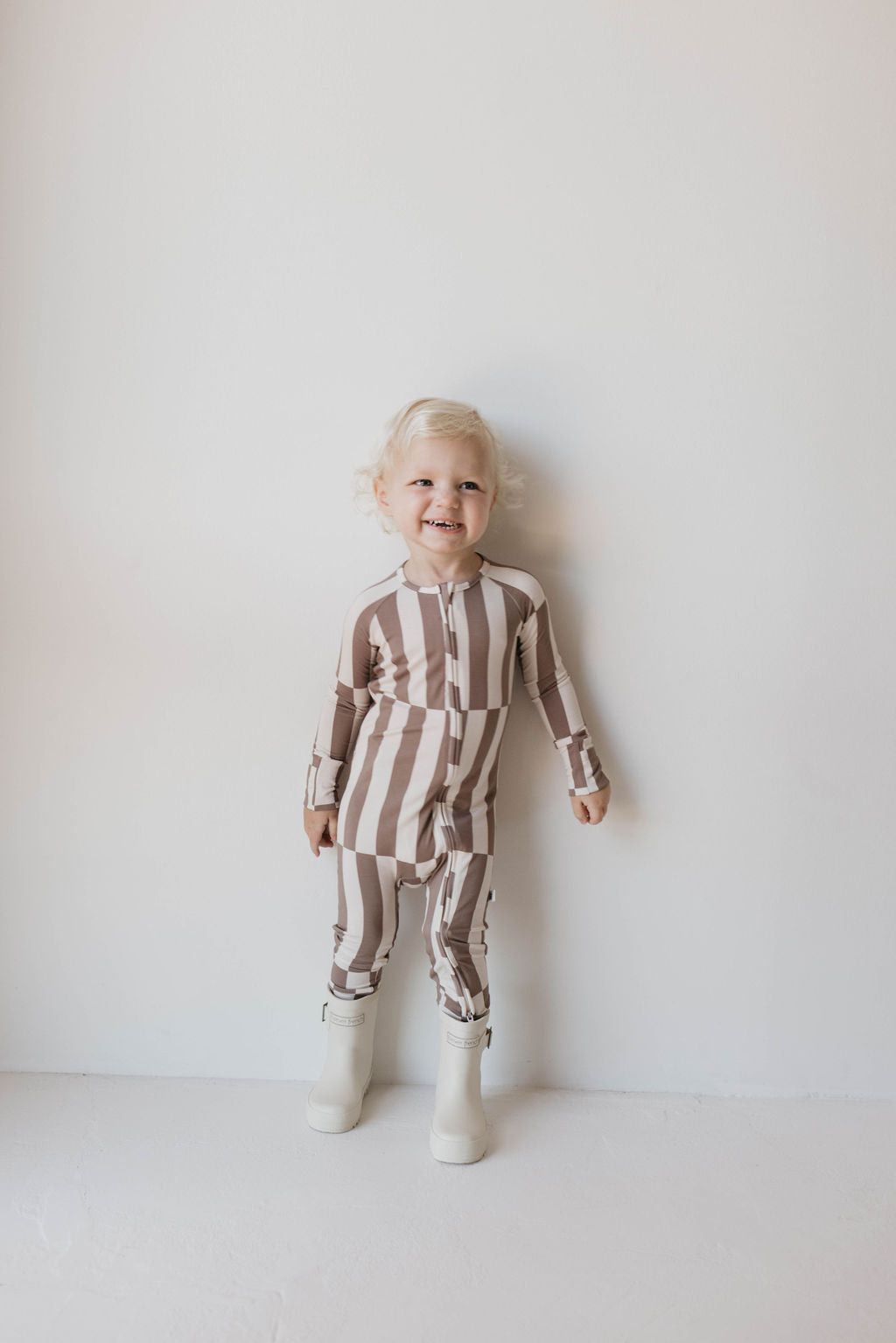 A smiling toddler with blonde hair stands against a plain white wall. The child is wearing a long-sleeve, striped outfit in beige and white from forever french baby, specifically the Bamboo Zip Pajamas | Tile Stack, along with white boots. These soft premium bamboo pajamas provide the ultimate comfort for your little one.