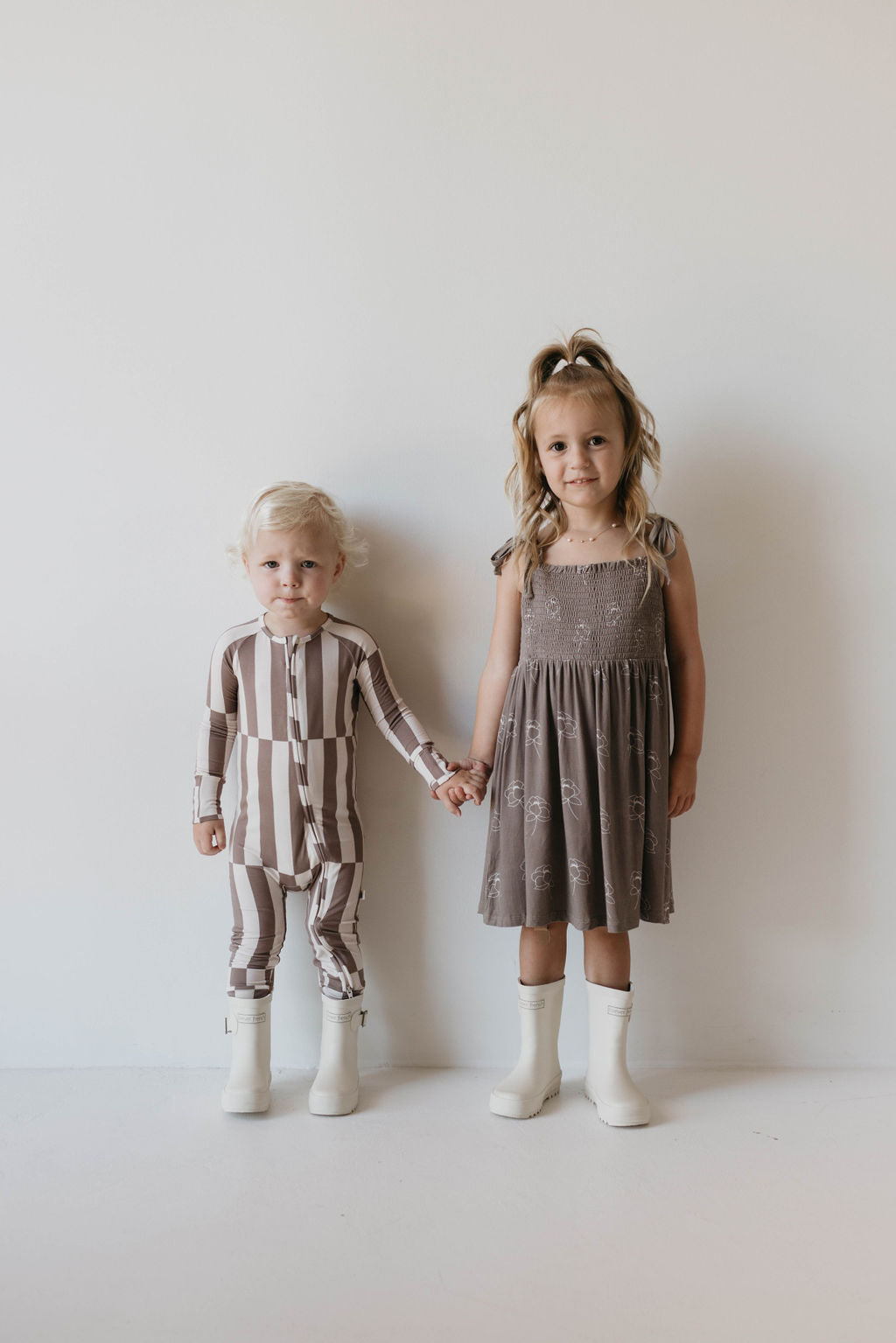 Two young children stand in front of a plain white background, holding hands. The child on the left has blonde hair, wears Bamboo Zip Pajamas in a Tile Stack pattern, and white boots. The child on the right has long hair in a ponytail, wears a forever french baby gray dress with a pattern, and white boots.