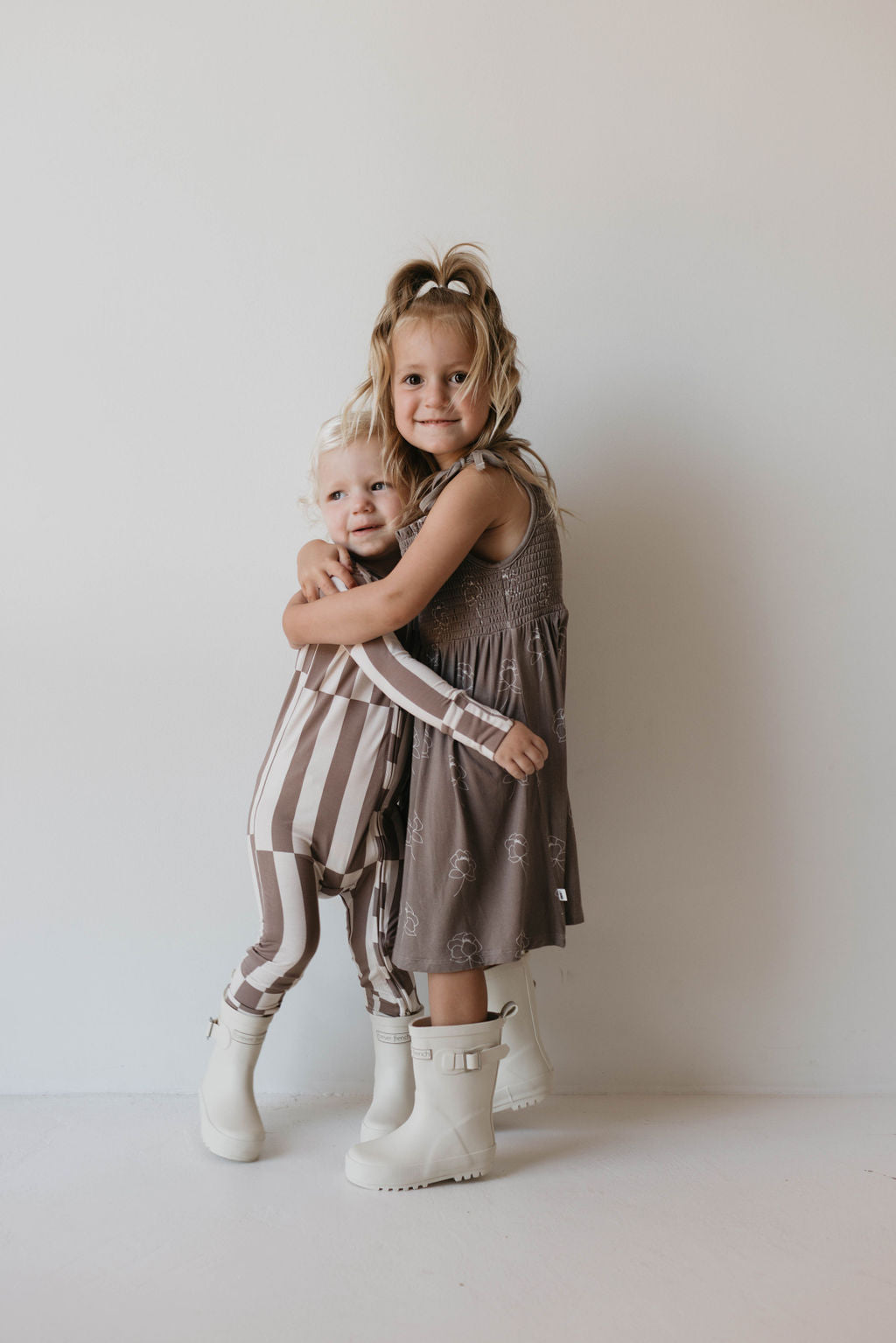 Two young children stand against a plain white wall. The older child, wearing a gray sleeveless dress and white boots, hugs the younger child, who is dressed in Forever French Baby's Bamboo Zip Pajamas in "Tile Stack" with matching white boots. Both children are smiling warmly at the camera.