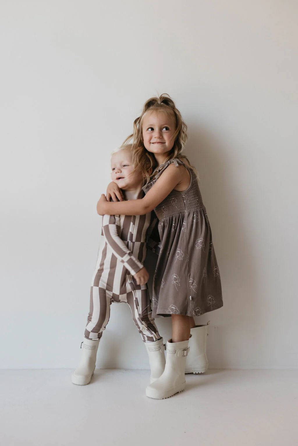 Two young children standing against a plain white background. The older child, in a brown dress with white patterns and ff Rain Boots in Cream by forever french baby, embraces the younger sibling, who is dressed in a striped brown and white outfit with matching all-weather kids footwear. Both are smiling warmly.