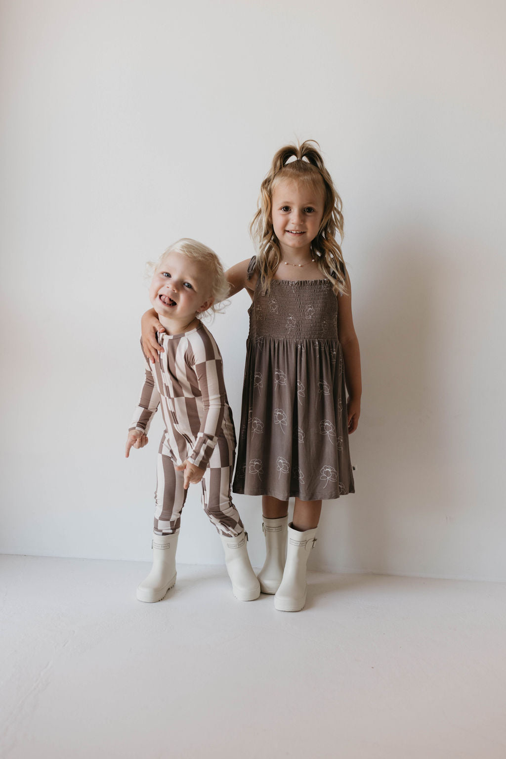 Two young children stand against a plain white background. One child, wearing a long-sleeve checkered outfit, smiles and leans into the other, who is clad in a Bamboo Tie Top Dress from forever french baby's Sweet Dreams Floral collection. Both are wearing white boots, and the older child has an arm around the younger one.