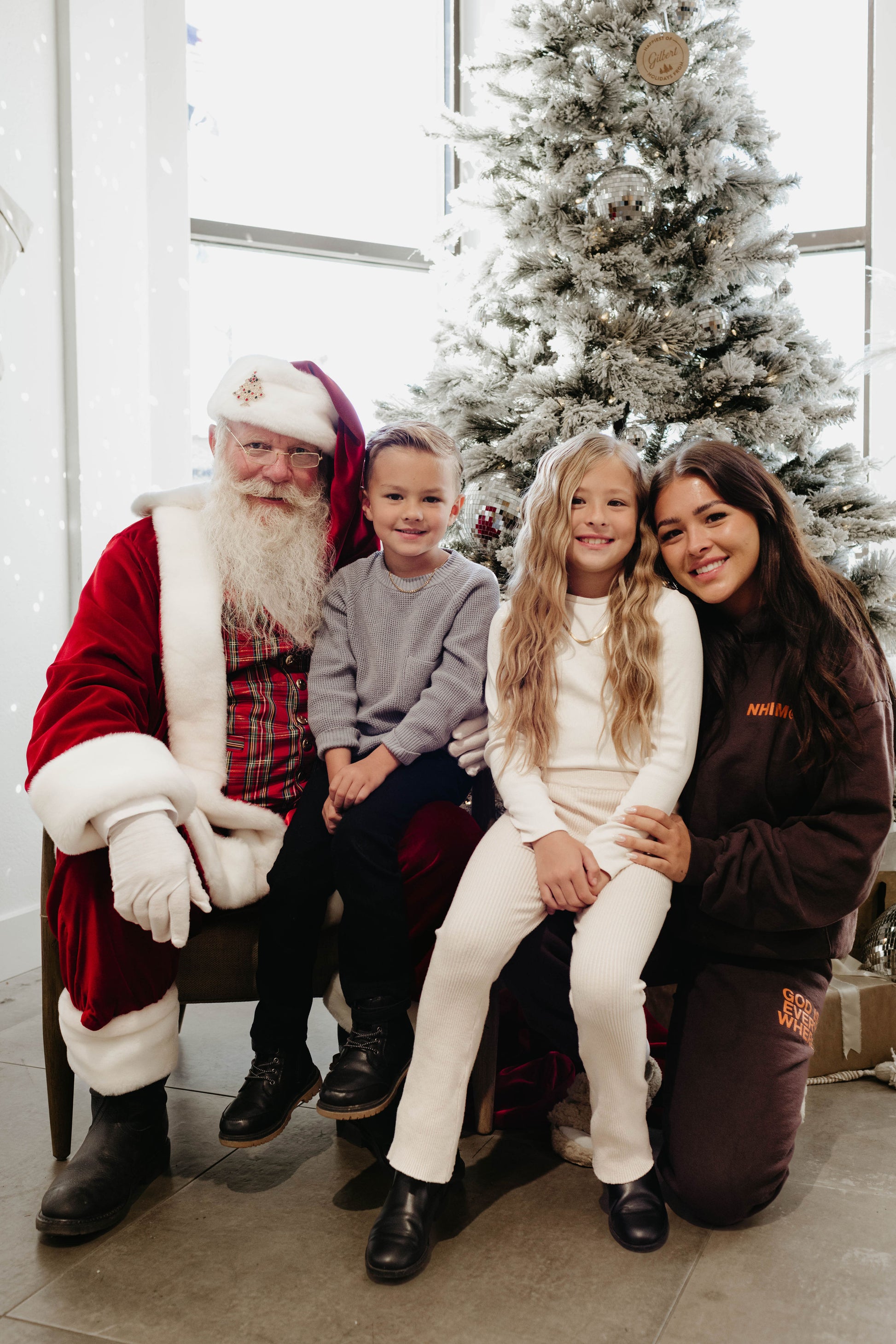 Santa Claus sits in a chair next to a beautifully decorated Christmas tree, with two children—a small boy and a girl—on his lap. A woman crouches beside them, smiling for the photo captured by Amanda Riley Photos as part of their Santa Photos | Forever French Baby x Amanda Riley Photos collection. Everyone appears happy and is enjoying the festive holiday moment indoors.