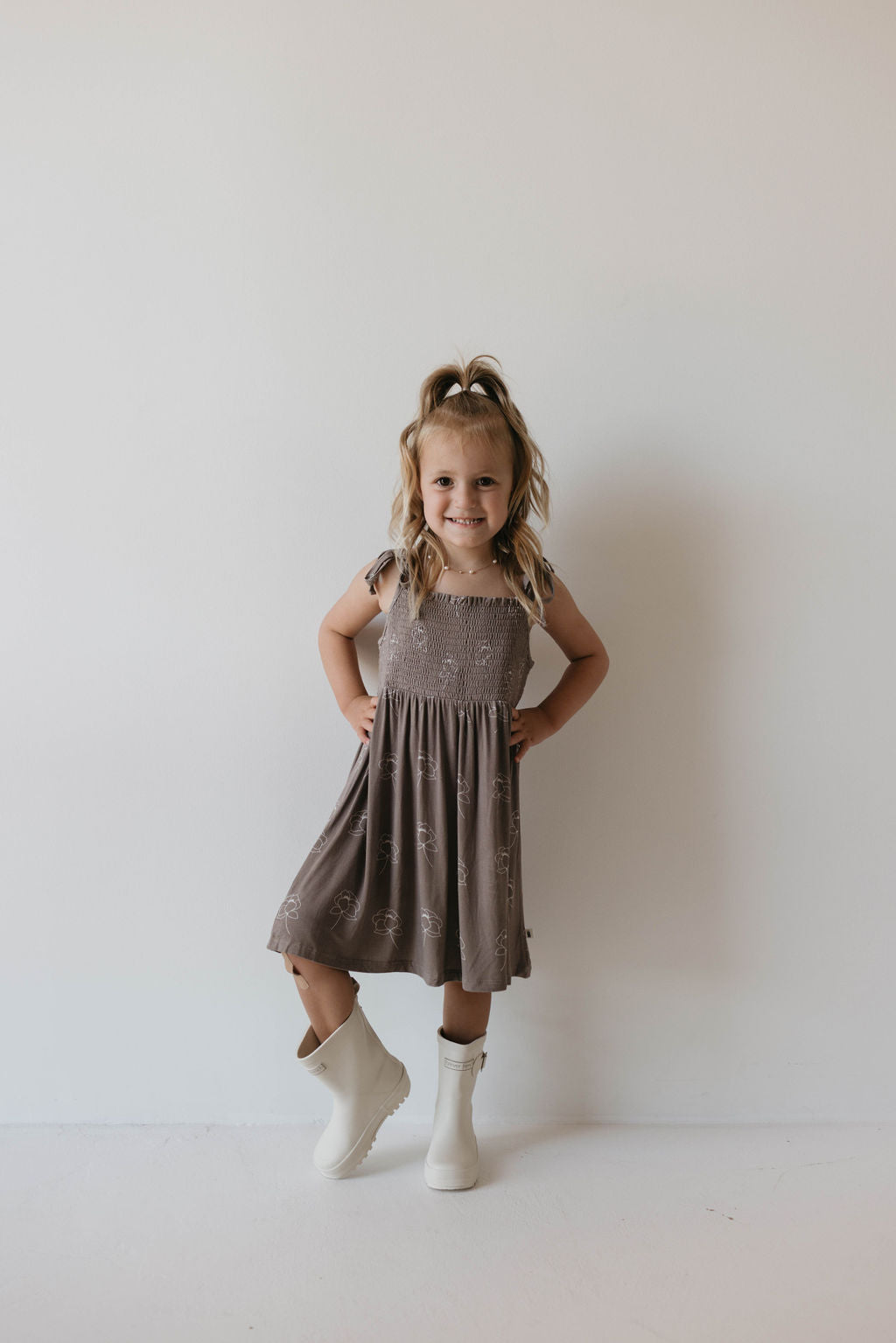 A young girl stands against a plain white wall, smiling. She has light brown hair styled in a half-up ponytail. She wears the knee-length Bamboo Tie Top Dress from the Sweet Dreams Floral collection by forever french baby, paired with short-sleeved white boots. Her hands rest confidently on her hips, radiating charm and confidence.