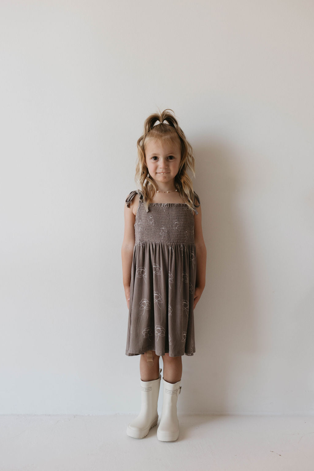 A little girl with light blonde hair stands against a white background, wearing the sleeveless Bamboo Tie Top Dress | Sweet Dreams Floral by forever french baby and white knee-high boots. Her hair is styled in a half-up ponytail with loose waves, and she has a neutral expression.