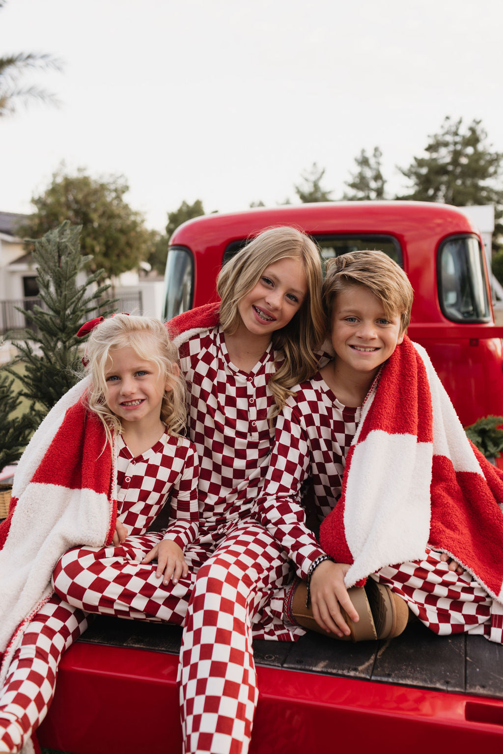 Three children wearing the Quinn bamboo two-piece pajamas by lolo webb, in a charming red and white checkered pattern, smile as they sit on the bed of a red pickup truck. They're cozily wrapped in a matching blanket, surrounded by a festive outdoor setting with trees providing the perfect backdrop.