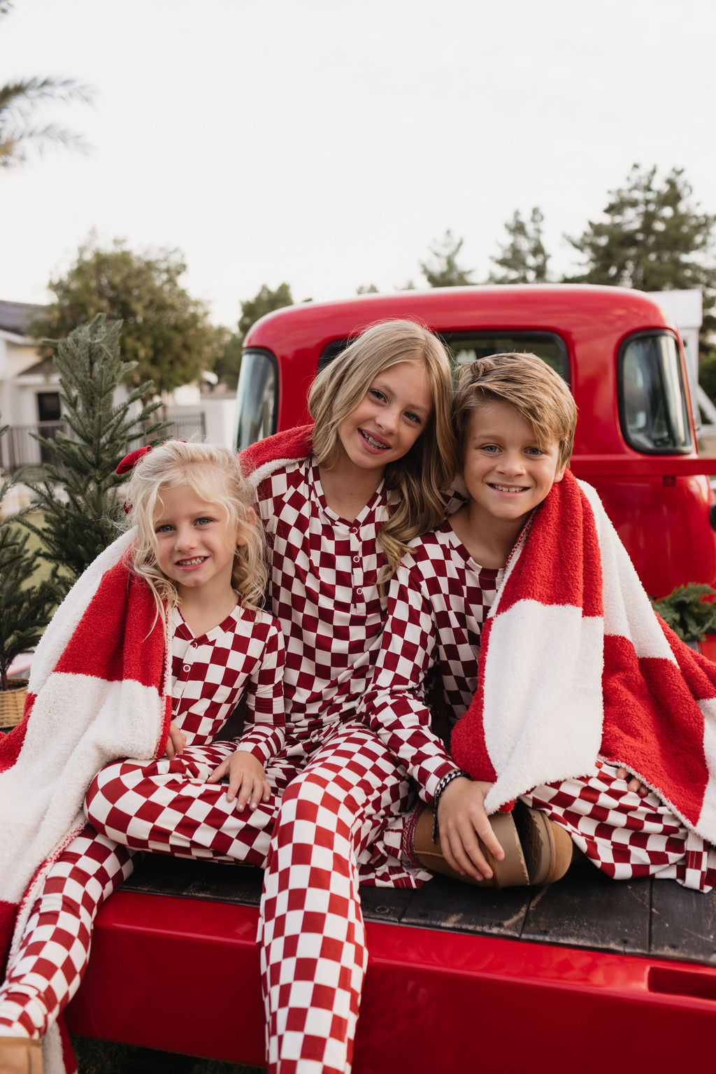 Three children wearing the Quinn bamboo two-piece pajamas by lolo webb, featuring a red and white checkered pattern, sit on the back of a vintage red truck. They are smiling and wrapped in a red and white blanket, with a small evergreen tree in the background.