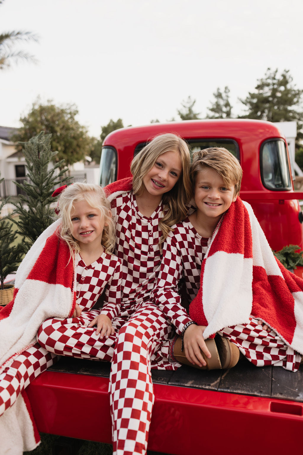 Three children wearing identical Quinn bamboo pajamas by lolo webb sit on the tailgate of a red truck. They are wrapped in a breathable red and white blanket, smiling, with trees and greenery in the background.