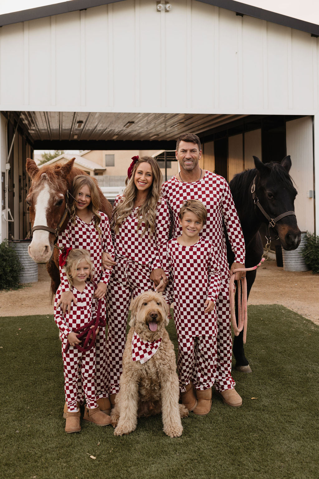 Clad in matching red and white checkered outfits made from the breathable fabric of lolo webb's Bamboo Two Piece Pajamas | the Quinn, a family of five joyfully poses with two horses and a dog beside a barn. The cheerful scene is enhanced as children, adults, and even the dog sport matching bandanas, creating an effortlessly coordinated look.