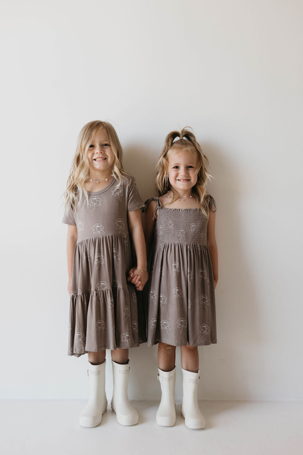 Two young girls with light hair stand side by side in matching Multi Tier Bamboo Dresses featuring the Sweet Dreams Floral pattern from forever french baby. They hold hands, each wearing white boots; one with straight hair and the other sporting a small ponytail tied with a bow. Both smile at the camera.