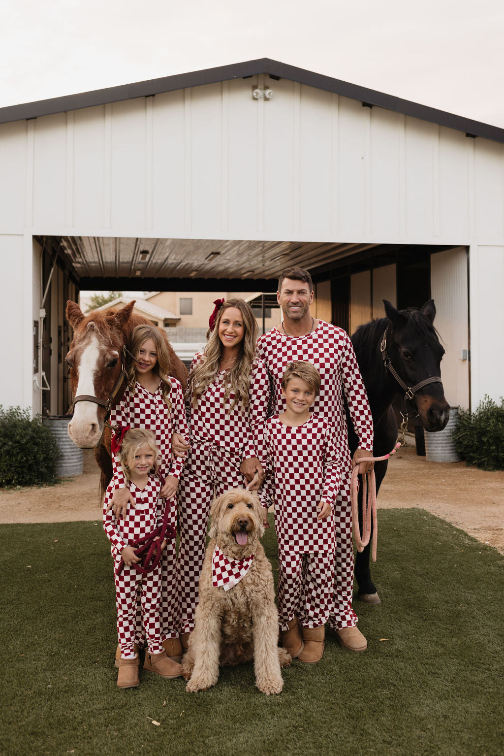 A family of five, dressed in matching Bamboo Two Piece Pajamas by lolo webb, stands in front of a barn with two horses and a large dog wearing a matching bandana. The scene is set outdoors on green grass.