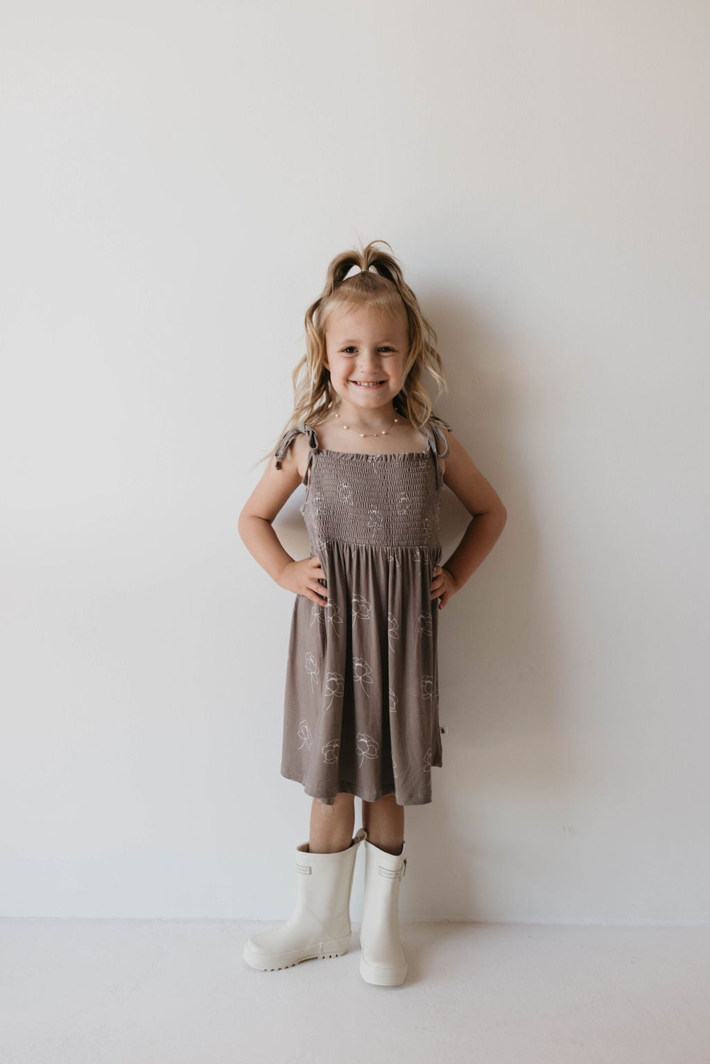 A young girl with light brown, shoulder-length hair, styled in a high ponytail, stands against a plain white wall. She smiles cheerfully with her hands on her hips while wearing the Multi Tier Bamboo Dress from forever french baby and white knee-high boots.