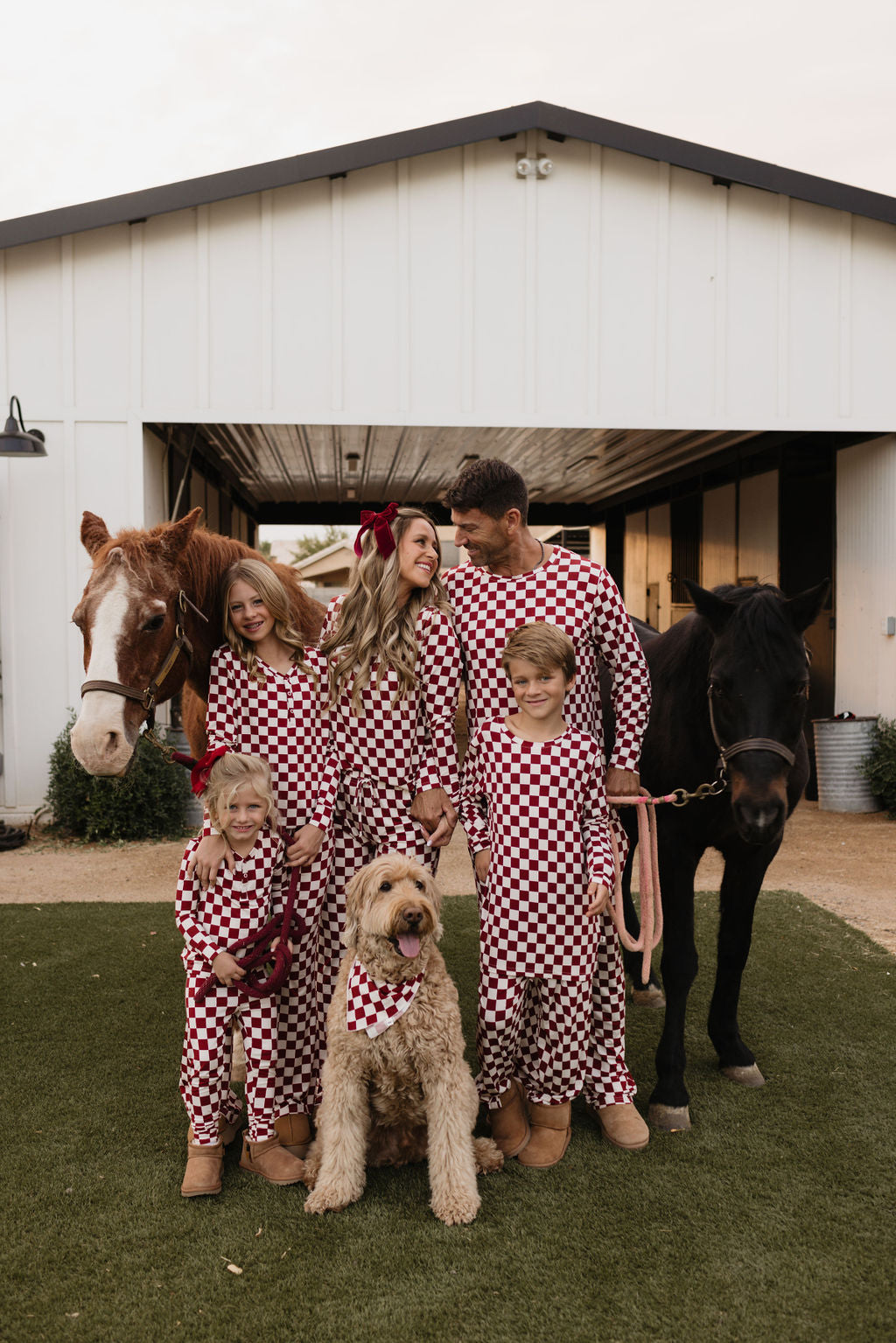 A family of five stands in front of a barn, dressed in matching Bamboo Two Piece Pajamas | the Quinn from lolo webb, featuring breathable fabric in a red and white checkered design. They are accompanied by two horses and a large dog. The adults and children are smiling, exuding a warm, cheerful atmosphere.