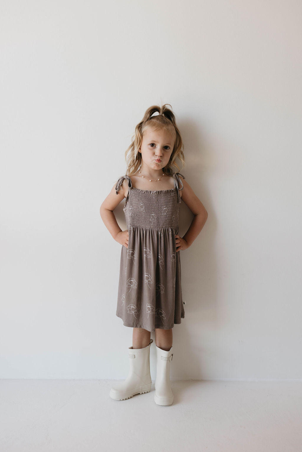 A young child with light brown hair stands against a plain white wall, staring directly at the camera with a serious expression. She is wearing a gray sleeveless "Multi Tier Bamboo Dress" featuring the "Sweet Dreams Floral" pattern from forever french baby, paired with white knee-high boots. Her hands rest on her hips.