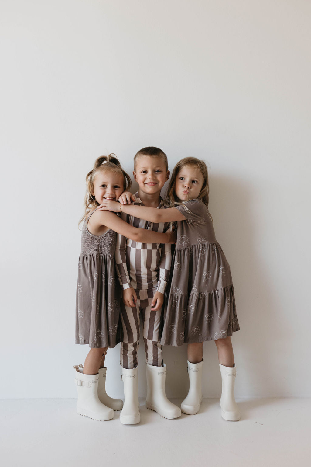 Three children stand against a light-colored wall, smiling and posing together. The two girls on the sides wear matching Sweet Dreams Floral Multi Tier Bamboo Dresses from forever french baby paired with white boots, while the boy in the middle wears a striped outfit. The children appear happy and close, embracing each other in their sweet dreams.