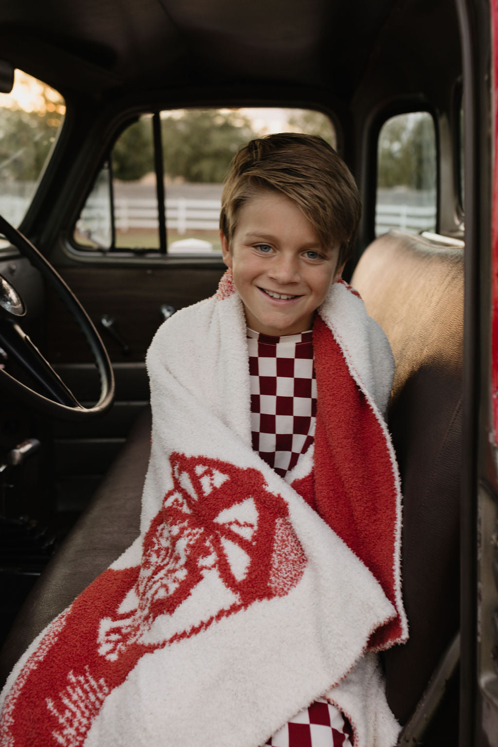 A young boy with short brown hair is comfortably seated in a vehicle, wrapped in a checkered red and white blanket, smiling. He wears the Pre-Teen Straight Leg Bamboo Pajamas from forever french baby, and sits on a dark bench seat. Through the window, a white fence and lush greenery can be seen outside.