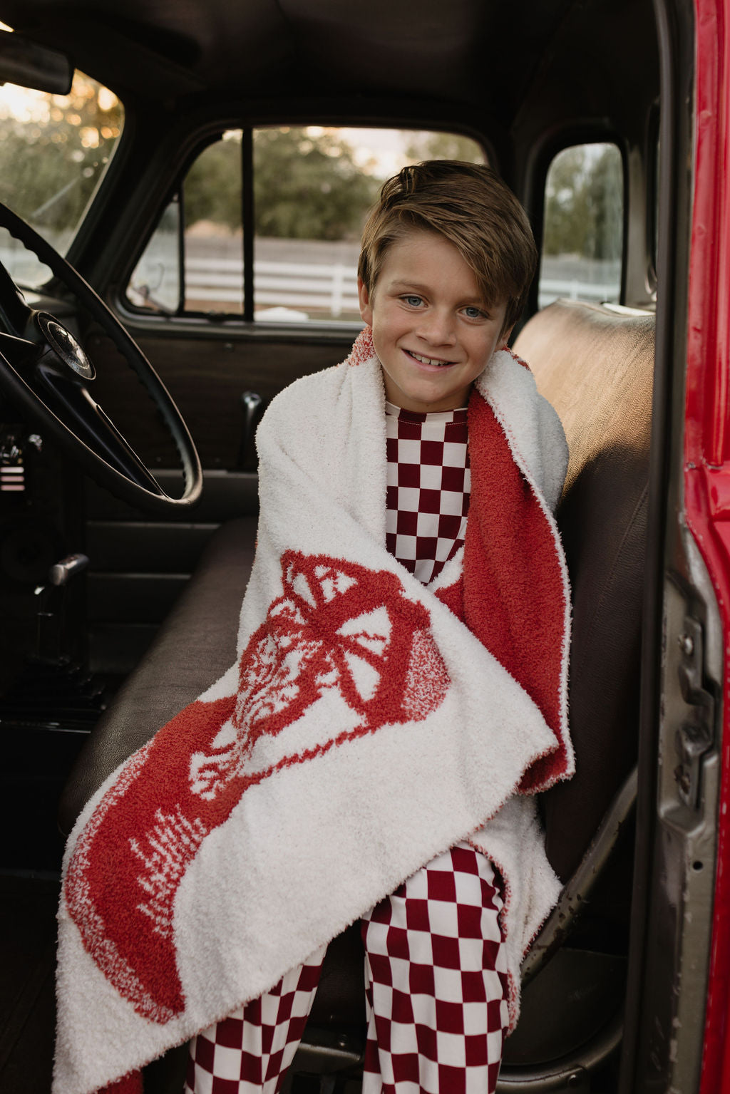 A smiling child sits inside a vehicle, wrapped in a white and red blanket, wearing the Pre-Teen Straight Leg Bamboo Pajamas called "the Quinn" from forever french baby. The pajamas boast a red and white checkered design made from breathable, hypo-allergenic fabric. The vehicle's interior is dark, with sunlight gently filtering through the window.