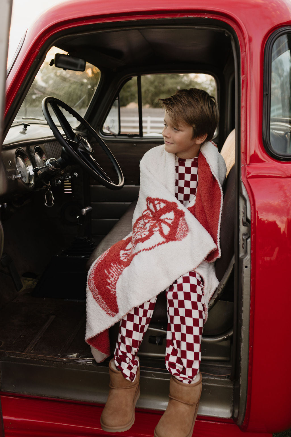 A young boy, wrapped in a red and white blanket, sits in the driver's seat of a vintage red truck. Wearing forever french baby’s Pre-Teen Straight Leg Bamboo Pajamas | the Quinn and brown boots, he looks content and cozy. The truck door is open, revealing the serene countryside outside.