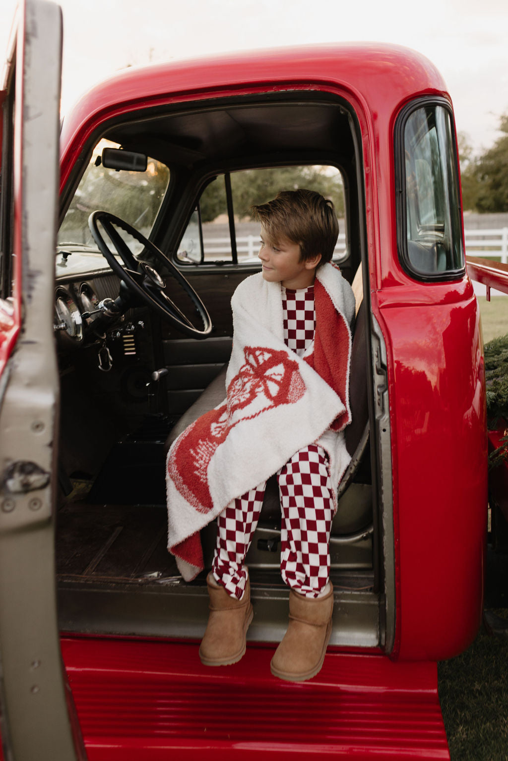 A young child sits in the driver's seat of a vintage red truck, wrapped in a white and red blanket. Dressed in forever french baby's breathable, hypo-allergenic Pre-Teen Straight Leg Bamboo Pajamas called "the Quinn," and wearing brown boots, they look out the open door with a relaxed expression.