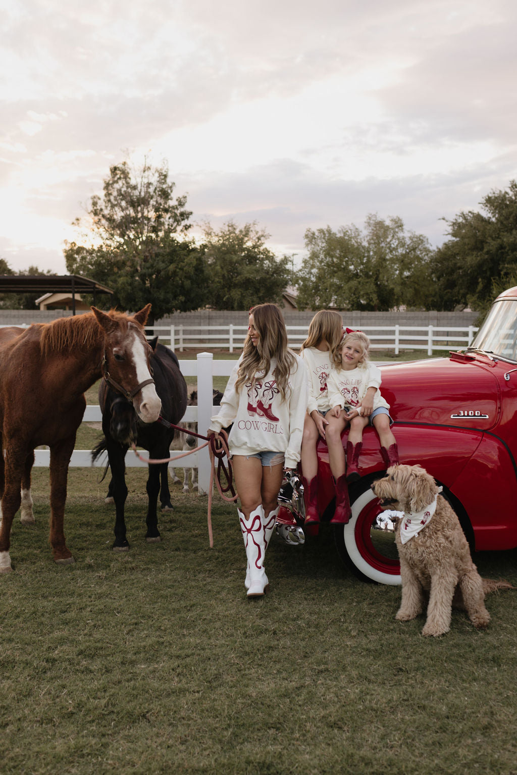 A woman in Western-style clothing stands in front of a red truck with two young girls wearing the Kids Crewneck | Long Live Cowgirls from lolo webb, all in cozy cotton outfits. Two horses stand nearby as a dog sits beside them, framed by a white fence and lush trees, creating a picturesque scene.