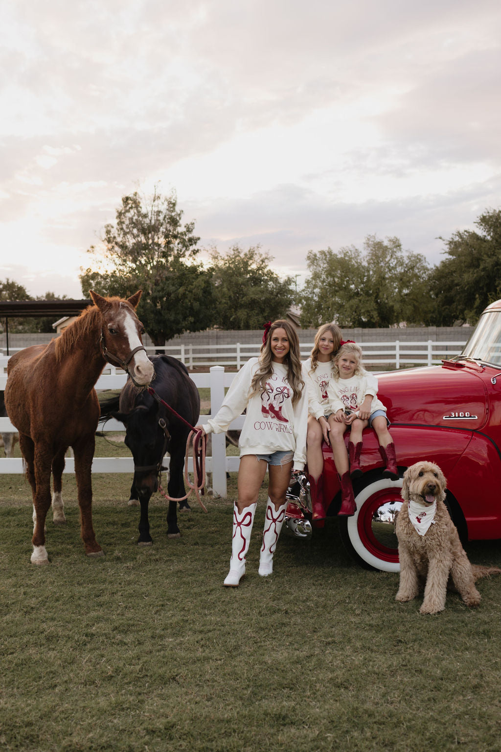 A woman and two children in matching cowboy-themed outfits stand near a red truck beside two horses. A fluffy dog lounges nearby. The scene, set on grassy ground with a white fence and trees, radiates warmth and charm, enhanced by their cozy Kids Crewneck | Long Live Cowgirls tops from lolo webb, made from cotton elastane.