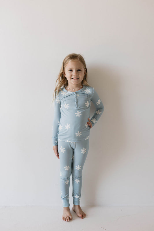 A young girl with long, light hair stands against a plain white background, wearing a matching blue outfit adorned with white flower patterns. She has one hand on her hip and is smiling at the camera. Her Daisy Blues outfit from forever french baby consists of the Bamboo Two Piece Pajamas, featuring a long-sleeve top and pants made from sustainable fabric.