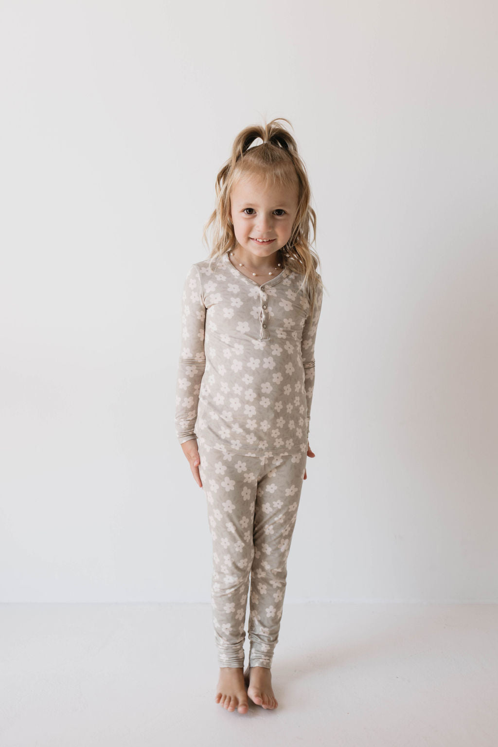 A young girl stands barefoot against a plain white background, smiling at the camera. She is wearing a matching "Bamboo Two Piece Pajamas | French Gray Floral" set from Forever French Baby, made from premium bamboo fabric with a light gray and white floral pattern. Her long blonde hair is tied up in a high ponytail.