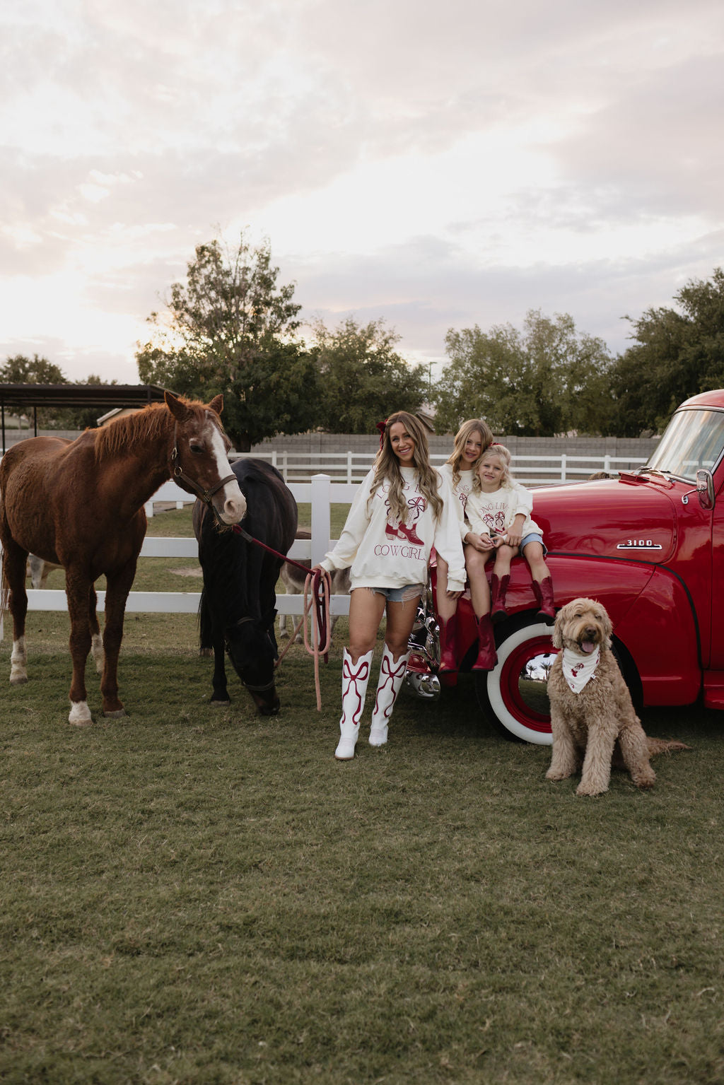 A woman wearing a cozy crewneck from lolo webb sits with two children on the hood of a vintage red truck, nestled in a grassy area. Nearby, a brown and black horse grazes quietly. A curly-haired dog lounges on the ground as clouds drift across the sky above a white fence in the background.