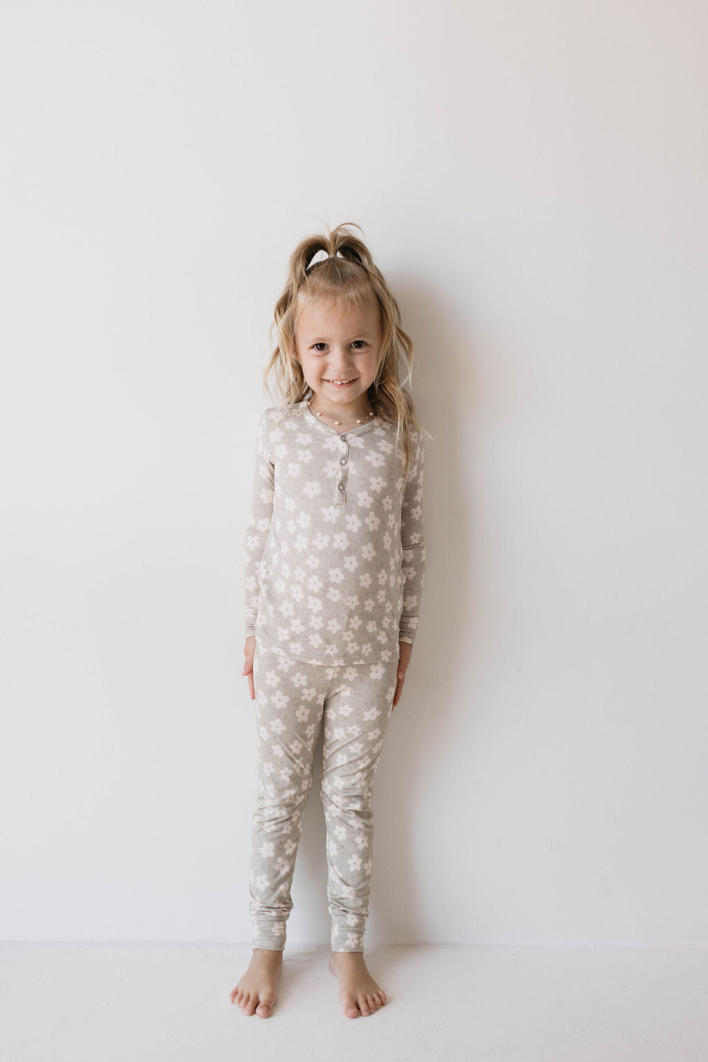 A young child with blonde hair tied in a high ponytail stands barefoot against a plain white background, wearing Forever French Baby's Bamboo Two Piece Pajamas in French Gray Floral. Made from premium bamboo fabric, the child smiles softly at the camera.