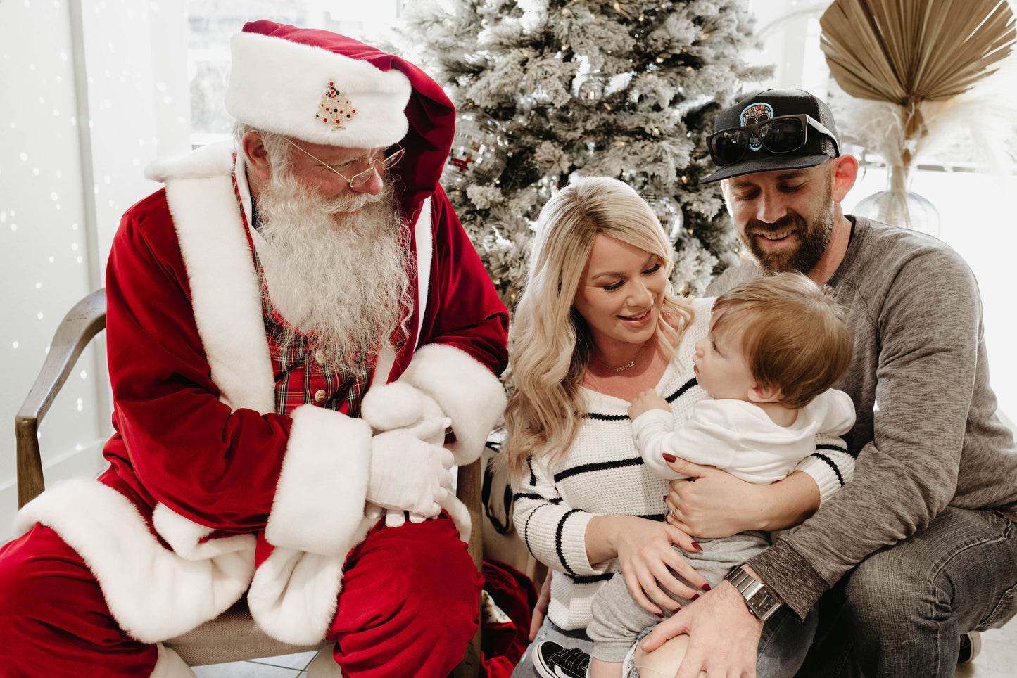 A family of three poses with Santa Claus in front of a beautifully adorned Christmas tree. The mother cradles a Forever French Baby, who gazes at Santa with curiosity. The father, sporting a hat and sunglasses, sits next to them, beaming with joy. This magical moment is immortalized by Amanda Riley Photos under the product name "Santa Photos | Forever French Baby x Amanda Riley Photos.