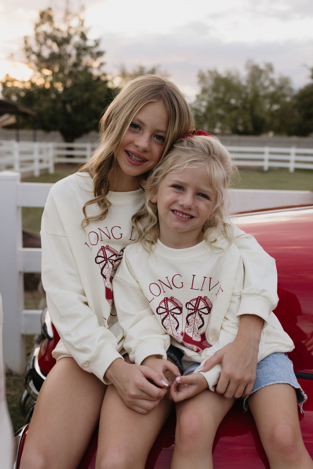 Two young girls sitting on a red car, smiling at the camera in their matching cream cozy crewneck sweatshirts from lolo webb, each featuring the "Long Live Cowgirls" design. Behind them, a white fence and trees frame the soft, cloudy sky.