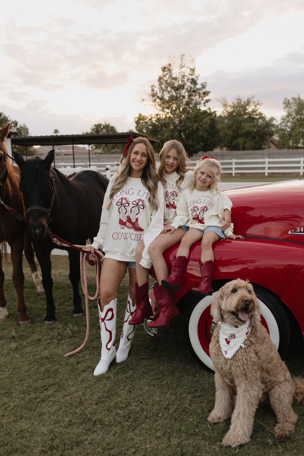 A woman and two young girls, dressed in coordinating bamboo viscose outfits featuring the "Long Live Cowgirls" print, stand near a red car. Accompanying them is a brown dog wearing the Bamboo Bandana from lolo webb’s "Cowgirl Christmas" collection and a horse. The scene is set outdoors against a backdrop of trees and a rustic fence.