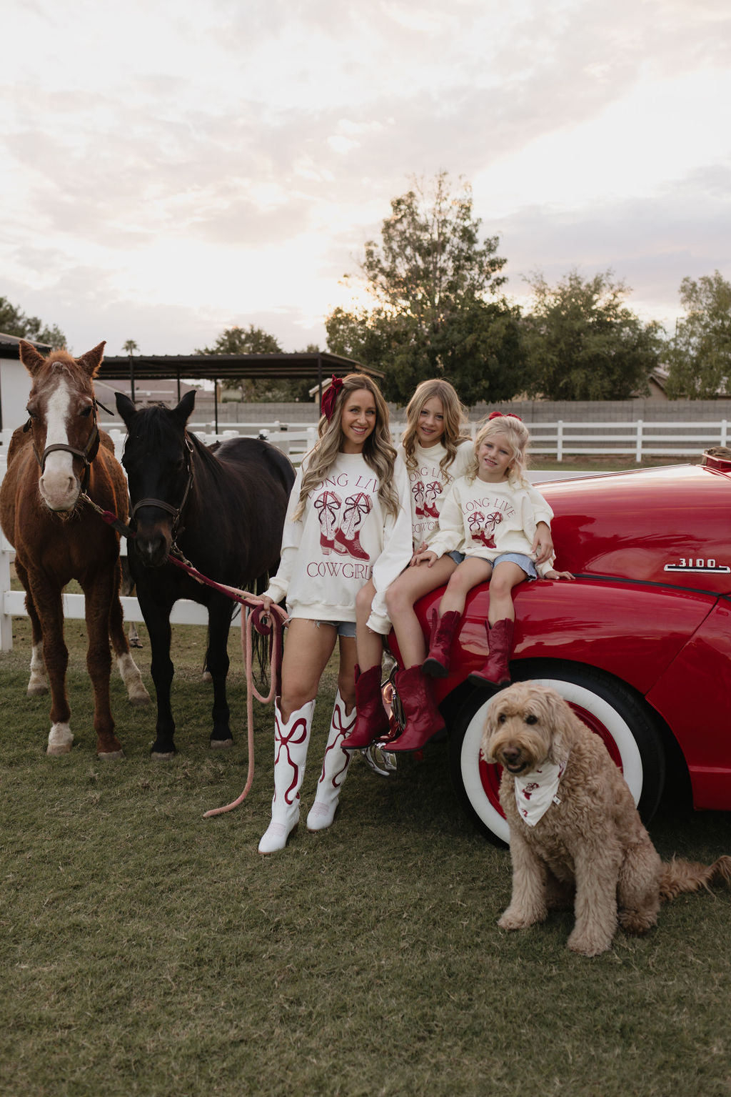 A woman stands with two children beside two horses and a red car, all wearing cozy crewnecks printed with "Long Live Cowgirls" from lolo webb. A dog sits at the front, sporting a bandana. The picturesque scene takes place in an open grassy area bordered by a white fence.