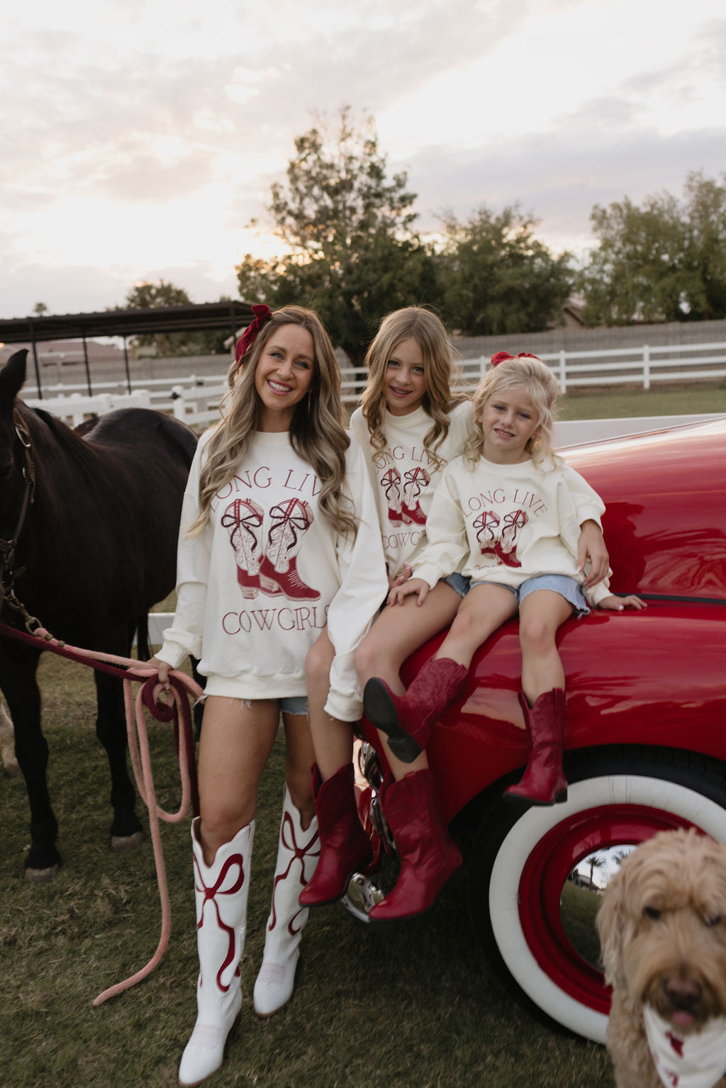 Three people wearing "Long Live Cowgirls" cotton shirts from lolo webb and red boots pose by a vintage red car. Two children sit on the hood, while a woman stands beside them holding a horse's reins. A dog is partially visible in the foreground, enhancing this charming holiday scene.