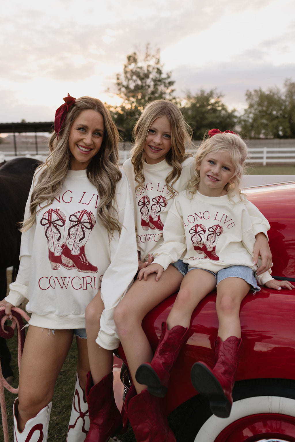 Three grinning girls clad in cozy Kids Crewneck shirts from lolo webb, each adorned with the "Long Live Cowgirls" design, and red boots are perched on a vintage red car. With bows adorning their hair, they sit outdoors beneath a cloudy sky, surrounded by trees.