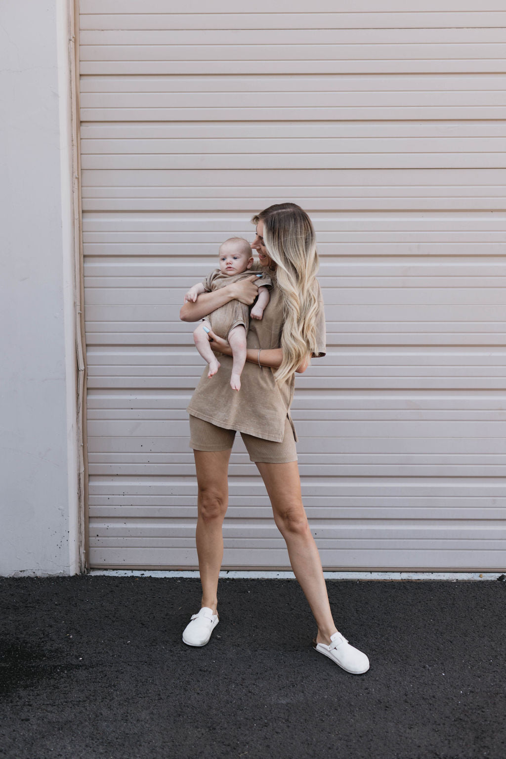 A person with long, blonde hair stands in front of a beige garage door, holding a baby from the brand forever french baby. Both are dressed in casual, neutral-toned outfits. The person is wearing the Women's Short Set in Vintage Washed Espresso and white sneakers while the baby rests calmly in their arms.