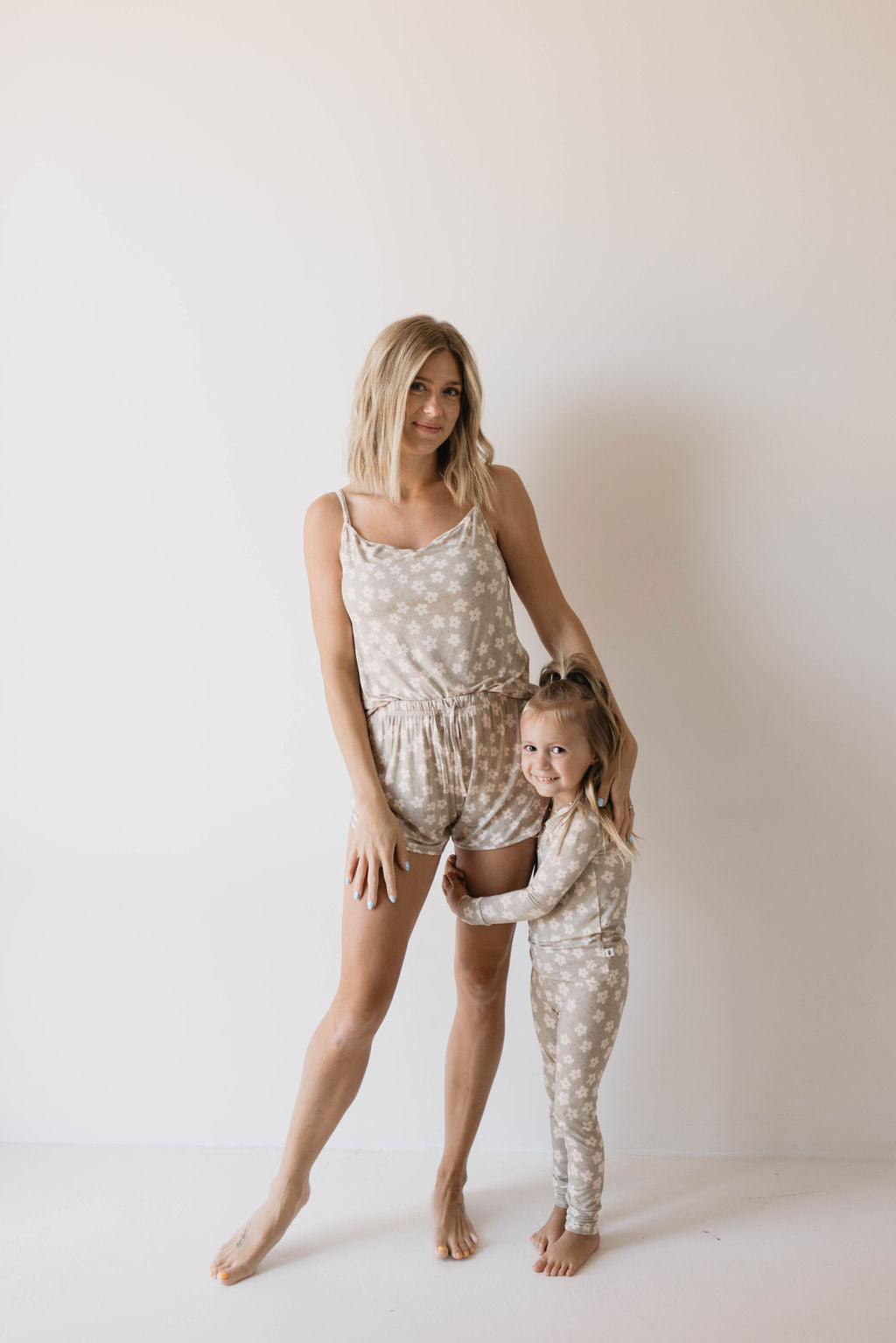 A woman and a young girl are standing against a plain white background. The woman is wearing a Cami Women's Bamboo Set in French Gray Floral by Forever French, while the girl is dressed in long-sleeved pajamas. Both outfits, made from soft bamboo clothing, feature a light beige color with a white star pattern. The girl is hugging the woman's leg.
