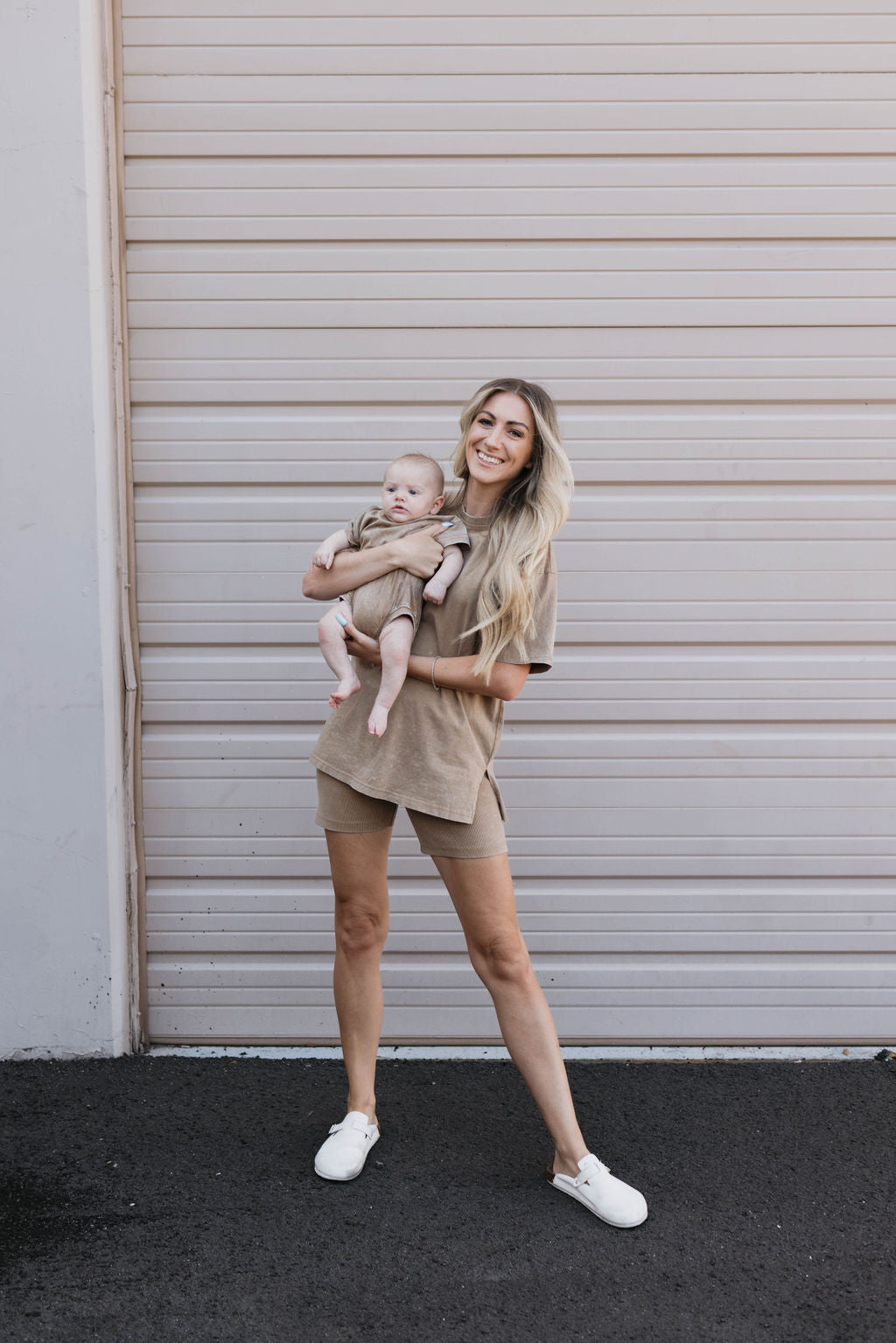 A woman with long blonde hair is wearing a Women's Short Set in vintage washed espresso, holding a smiling baby dressed in a forever french baby outfit. They are standing in front of a beige garage door on a paved surface, both appearing happy and relaxed.