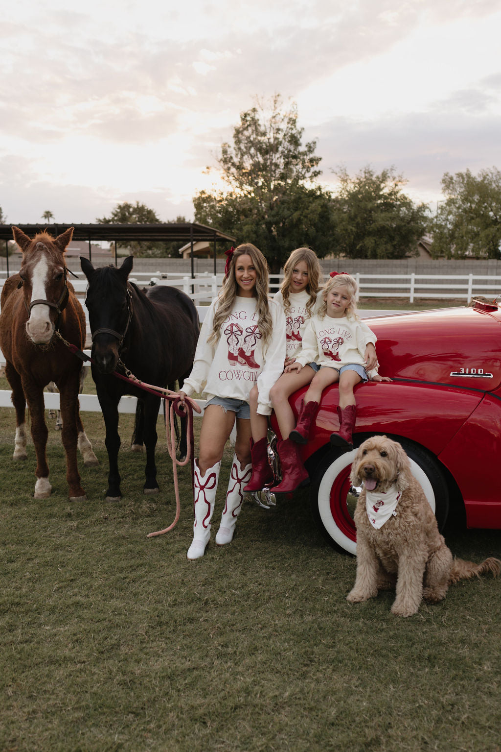 A woman and two children, dressed in matching bamboo viscose outfits with cursive designs, stand beside a vintage red car, two horses, and a goldendoodle wearing a lolo webb Bamboo Bandana in the Cowgirl Christmas style. They are outdoors on a grassy area with trees and a fence in the background.